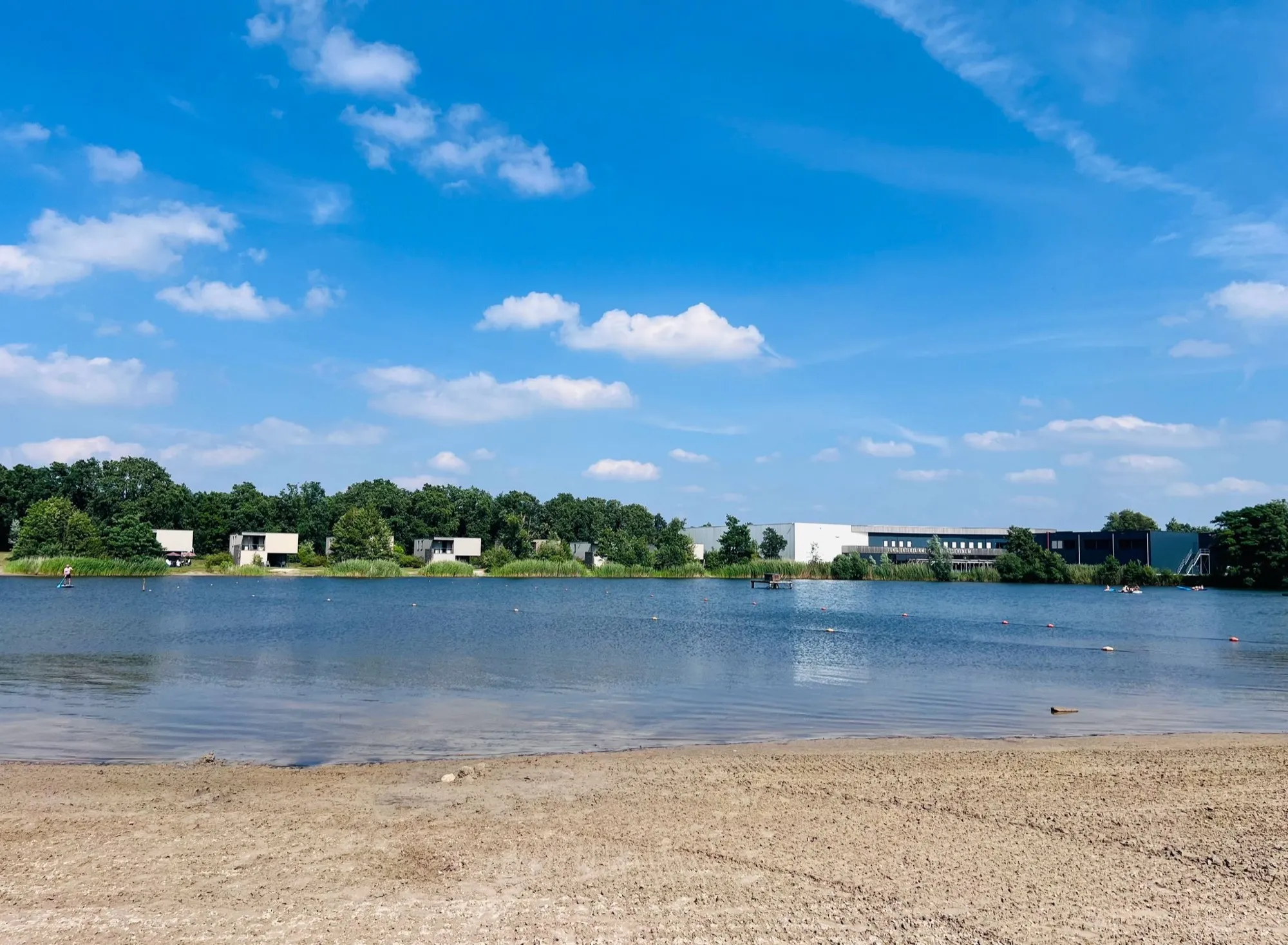 Sandstrand am Badesee des Ferienparks De Schatberg mit Blick über das Wasser auf Bäume und Parkgebäude am gegenüberliegenden Ufer