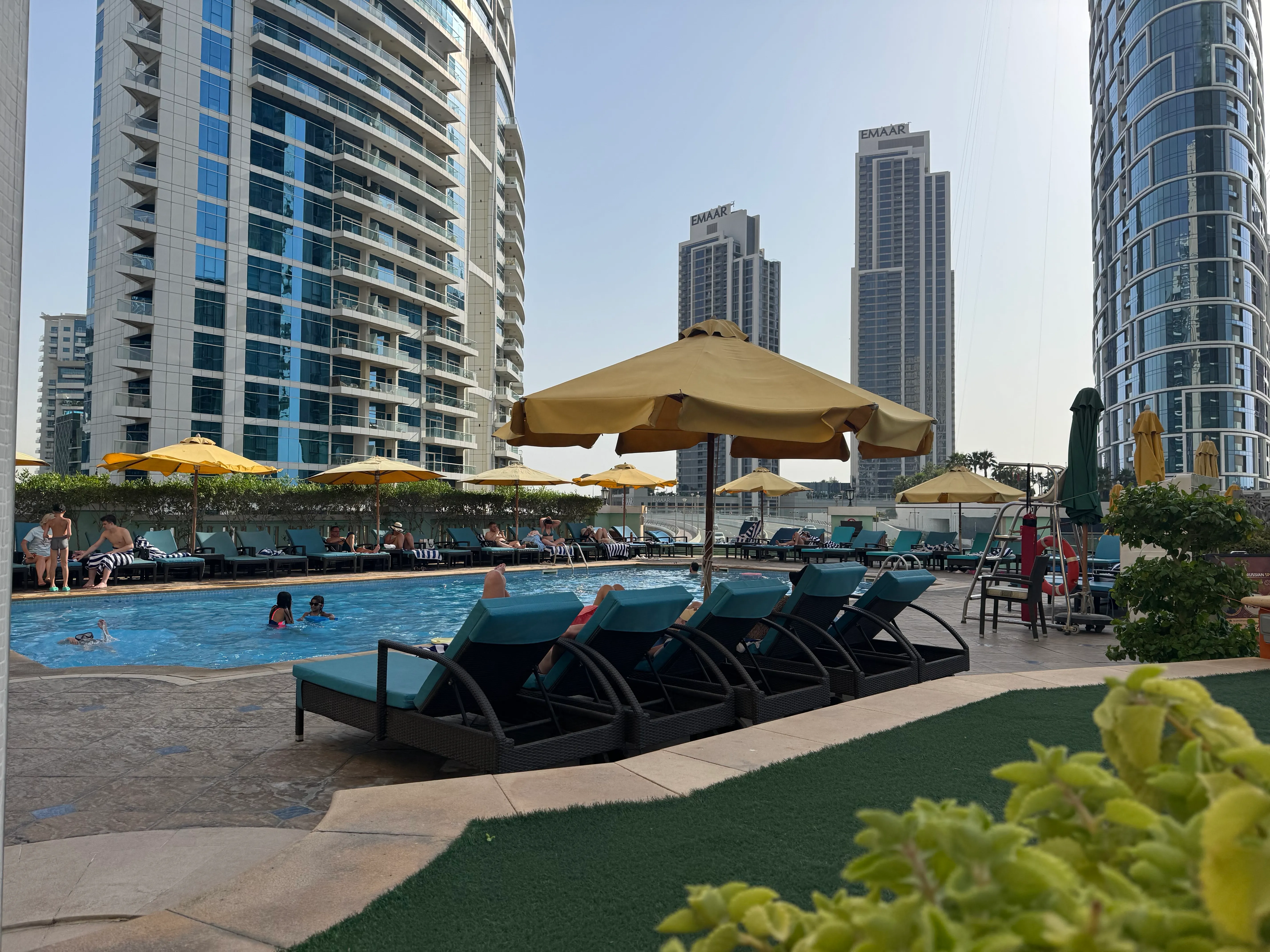 Poolbereich auf der Plaza des Ramada by Wyndham Dubai JBR mit Sonnenliegen und Hochhaus-Skyline