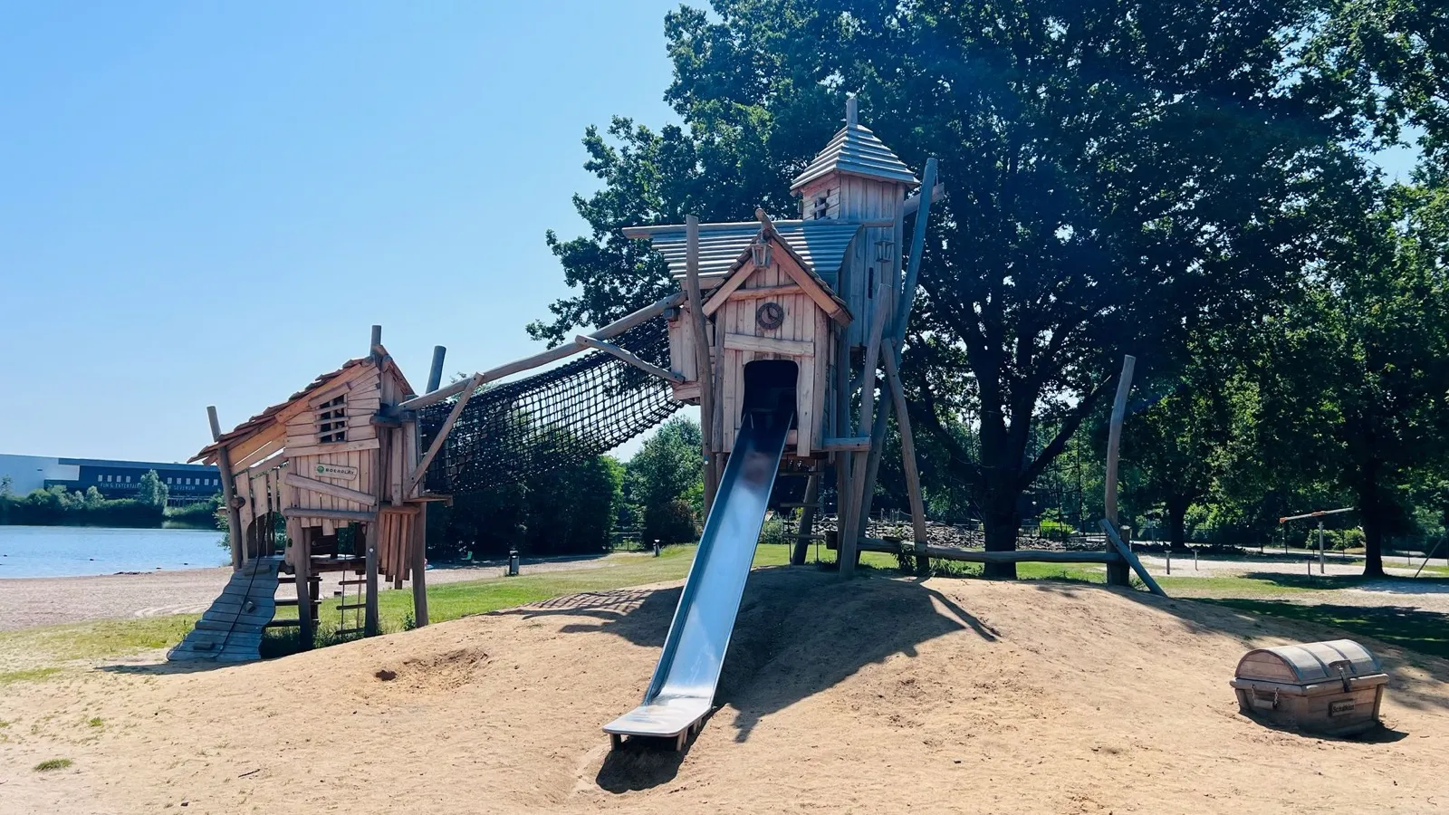 Piraten-Spielplatz aus Holztürmen mit langer Metallrutsche im Sand, direkt am Badesee-Strand