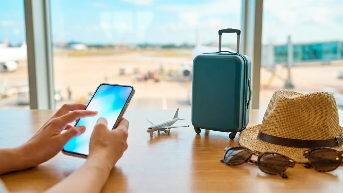 Symbolbild: Hände buchen am Smartphone Gepäck nach — Koffer und Mini-Flugzeug auf einem Holztisch